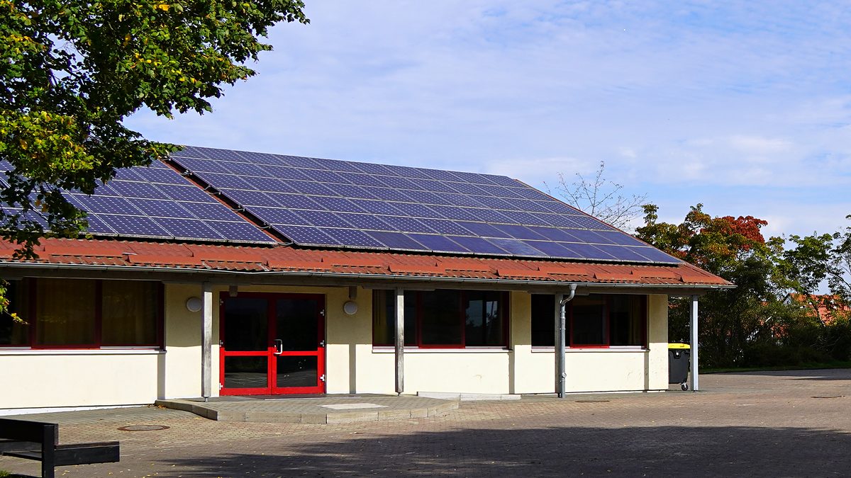 Der Pavillon mit Solaranlage auf dem Dach und roter Eingangstür, links ein Baum