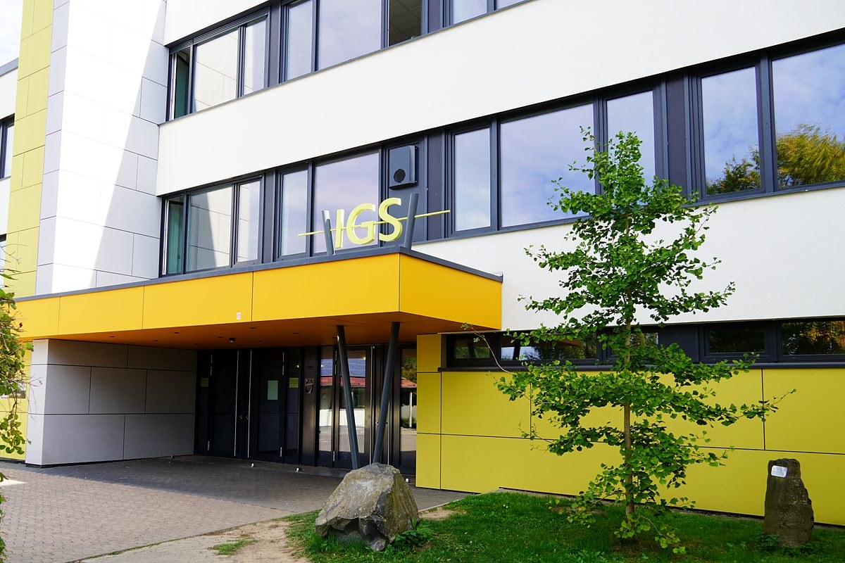 Haupteingang der IGS Guxhagen mit gelbem Vordach und IGS-Schriftzug, davor ein junger Baum und ein Findling auf dem Rasen