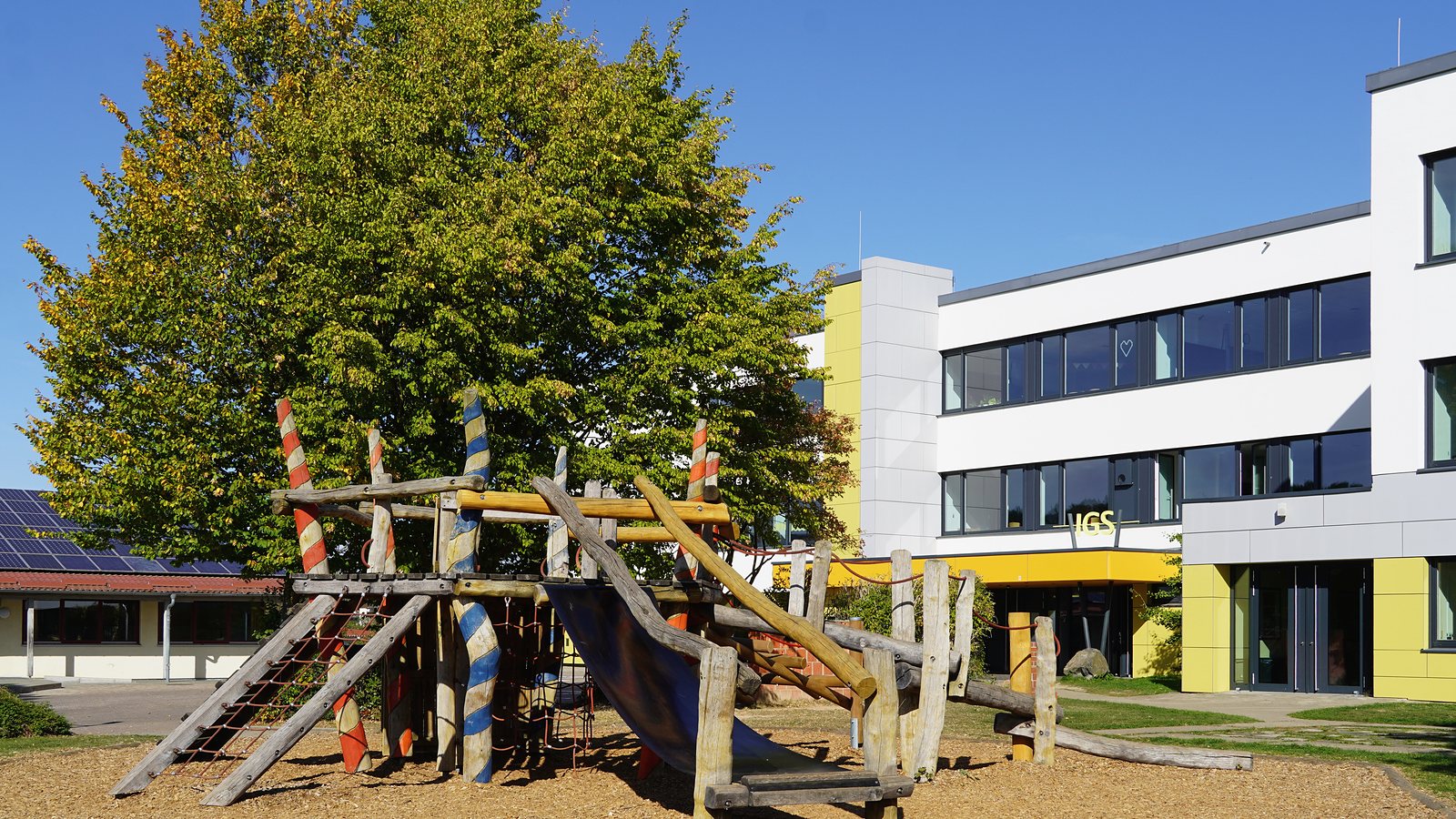 Schulhof der IGS Guxhagen im Herbst: Klettergerüst, bunte Bäume und das Hauptgebäude mit IGS-Schriftzug