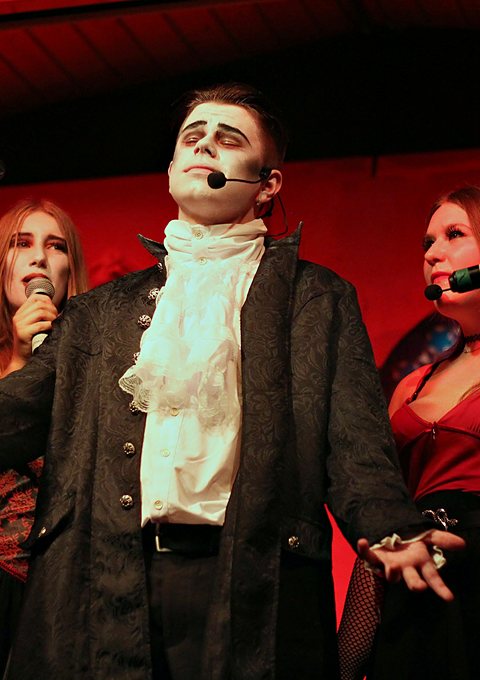 Szene aus dem Musical 2023: ein bleich geschminkter Graf mit Rüschenhemd vor rot beleuchteten Darstellerinnen