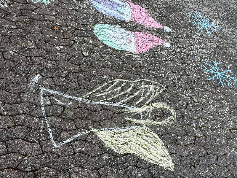Kreidebild auf dem Schulhof: ein Engel, darüber zwei Weihnachtsmützen und Schneeflocken