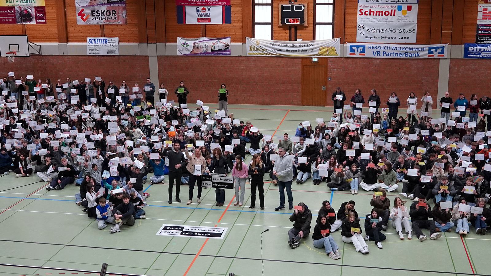 Die Schulgemeinde der IGS sitzt in der Sporthalle und hält selbst gestaltete Schilder hoch, vorne das Schild „Schule ohne Rassismus – Schule mit Courage“